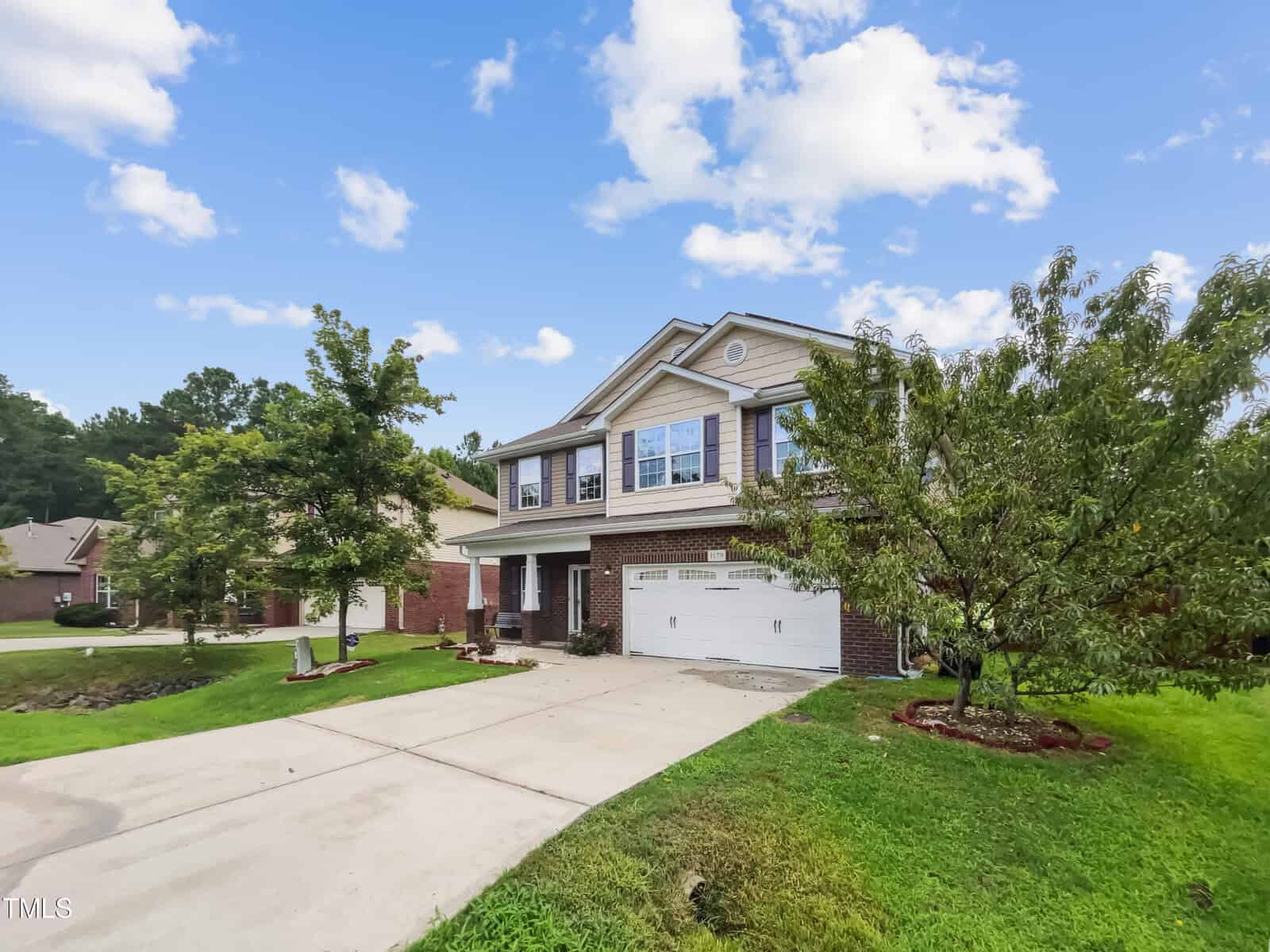 The two-story suburban house at 1179 Ridgeland Drive in Creedmoor features a brick and beige siding exterior, a white garage door, and a welcoming front porch. A driveway stretches to the street, surrounded by trees and a small lawn. Nearby houses dot the landscape under a partly cloudy sky.