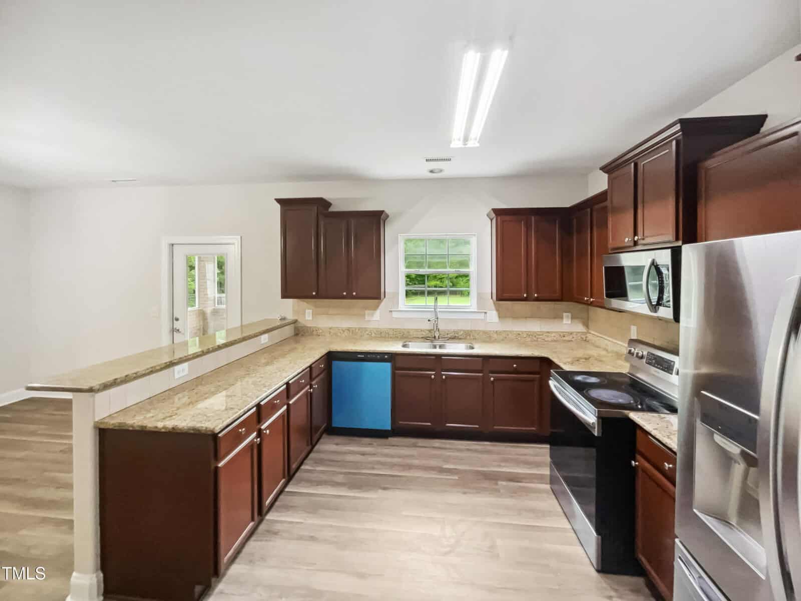 A kitchen with dark wooden cabinets, stainless steel appliances, and light brown granite countertops. There's a large window above the sink, a blue dishwasher, and an open layout leading to another room. The floor is wood.