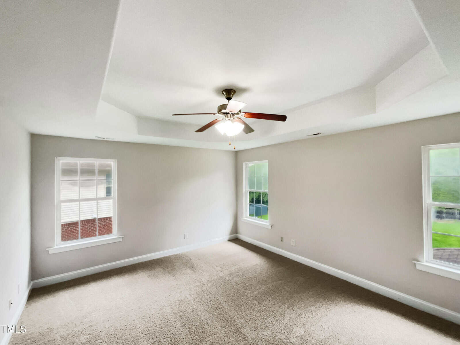 Empty room with beige carpet and light gray walls, featuring a white ceiling with a recessed design. A ceiling fan with five wooden blades and a light fixture hangs in the center. Two windows on adjacent walls let in natural light.