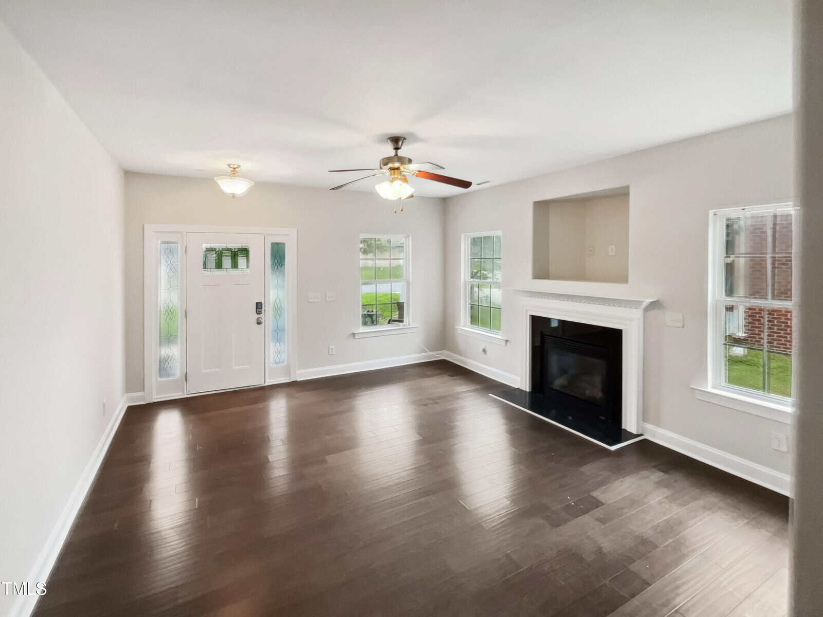 A living room with hardwood floors, a ceiling fan, and recessed lighting. It features a fireplace with a mantle, two large windows, and a white front door with frosted glass panels. The walls are painted light gray.