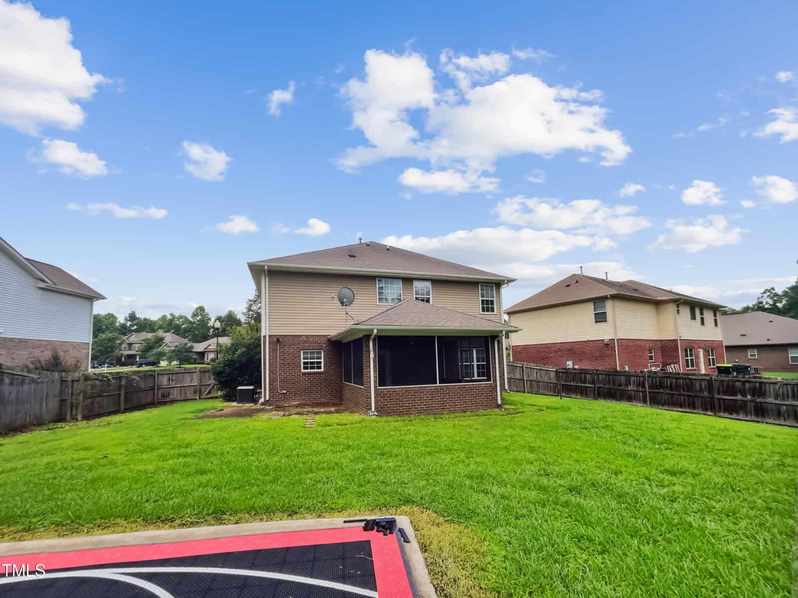A two-story brick house with a fenced backyard and well-maintained lawn under a partly cloudy sky. A small concrete area with a red and black design is visible in the foreground.
