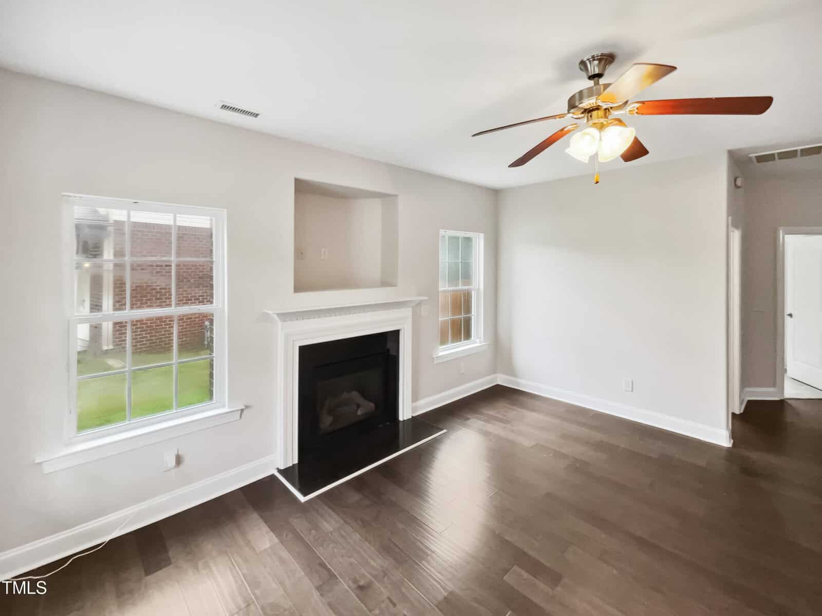 A living room with light-colored walls and a dark hardwood floor. It features a white fireplace, a ceiling fan with lights, and two windows on either side of the fireplace, allowing natural light into the room.