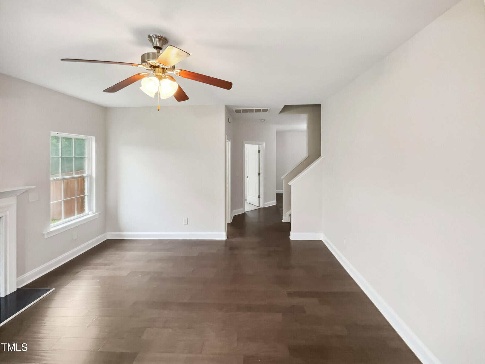A clean, unfurnished living room with wooden floors, a ceiling fan with lights, a window on the left, and a white fireplace. The room leads to a hallway and staircase on the right. Walls and ceiling are painted white.