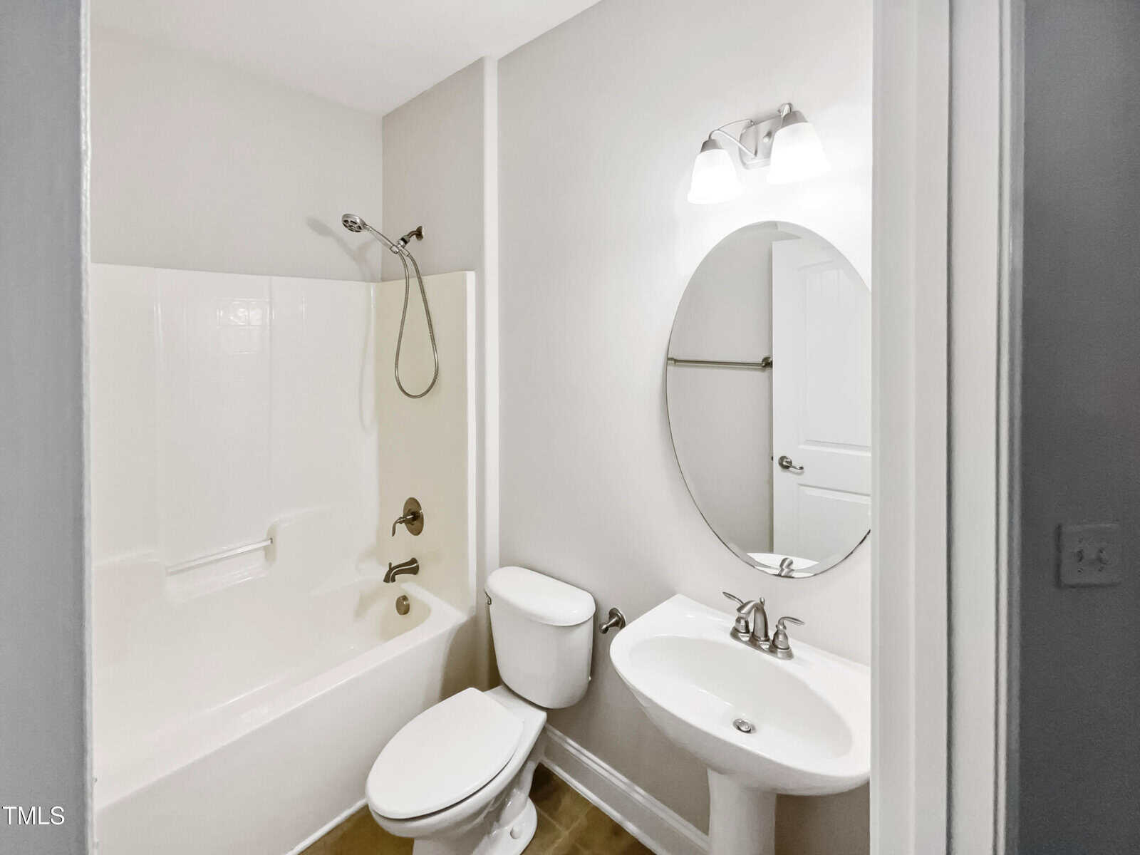 A small bathroom features a white bathtub with a showerhead, a toilet, and a pedestal sink. An oval mirror hangs above the sink, and a light fixture with two bulbs is positioned above the mirror. Walls and fixtures are white, creating a clean appearance.