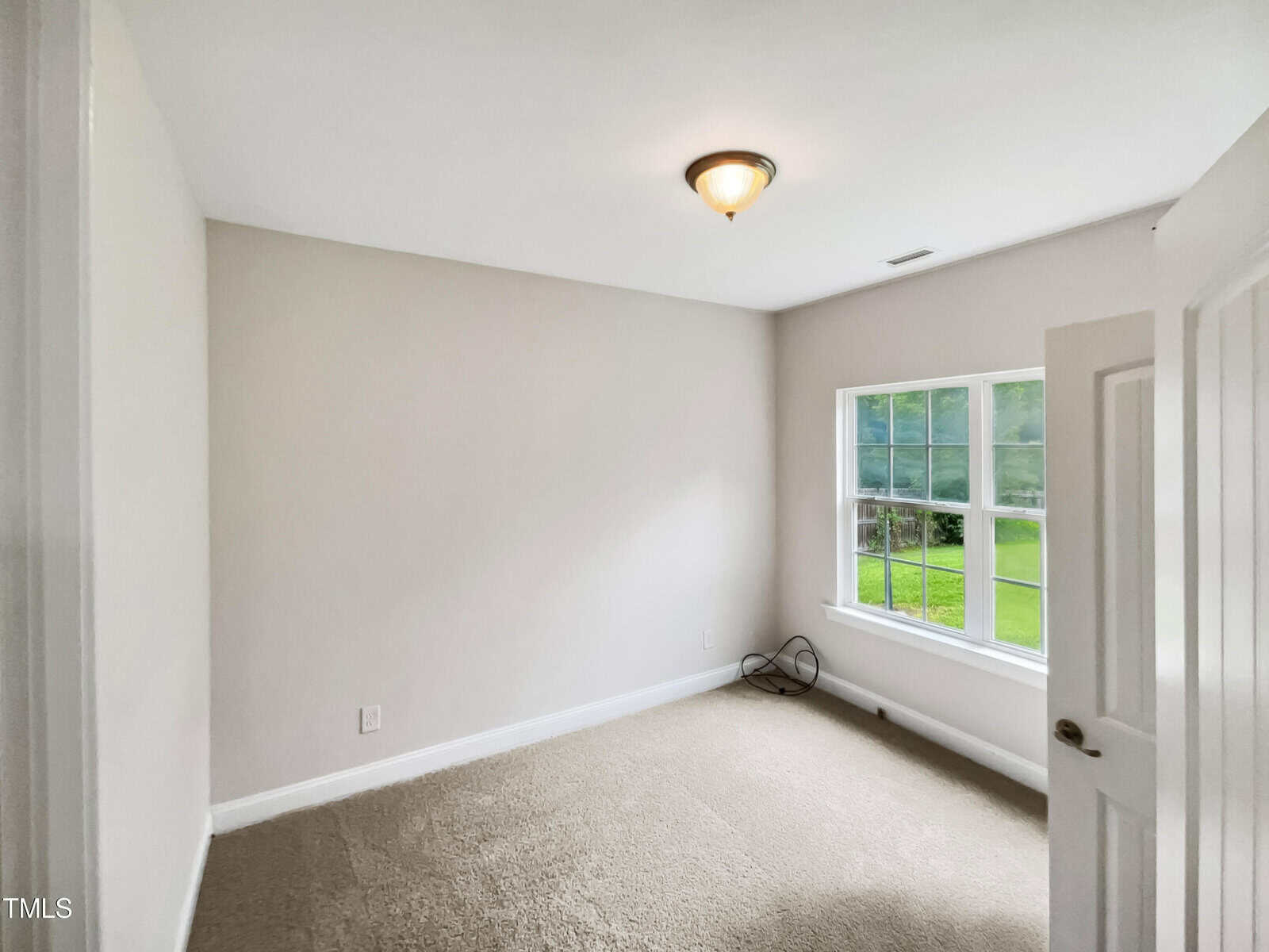 A small, empty room with beige carpet and white walls. A single ceiling light is on. Sunlight enters through a window on the right, and an open white door is on the left. A cable is coiled near the wall below the window.