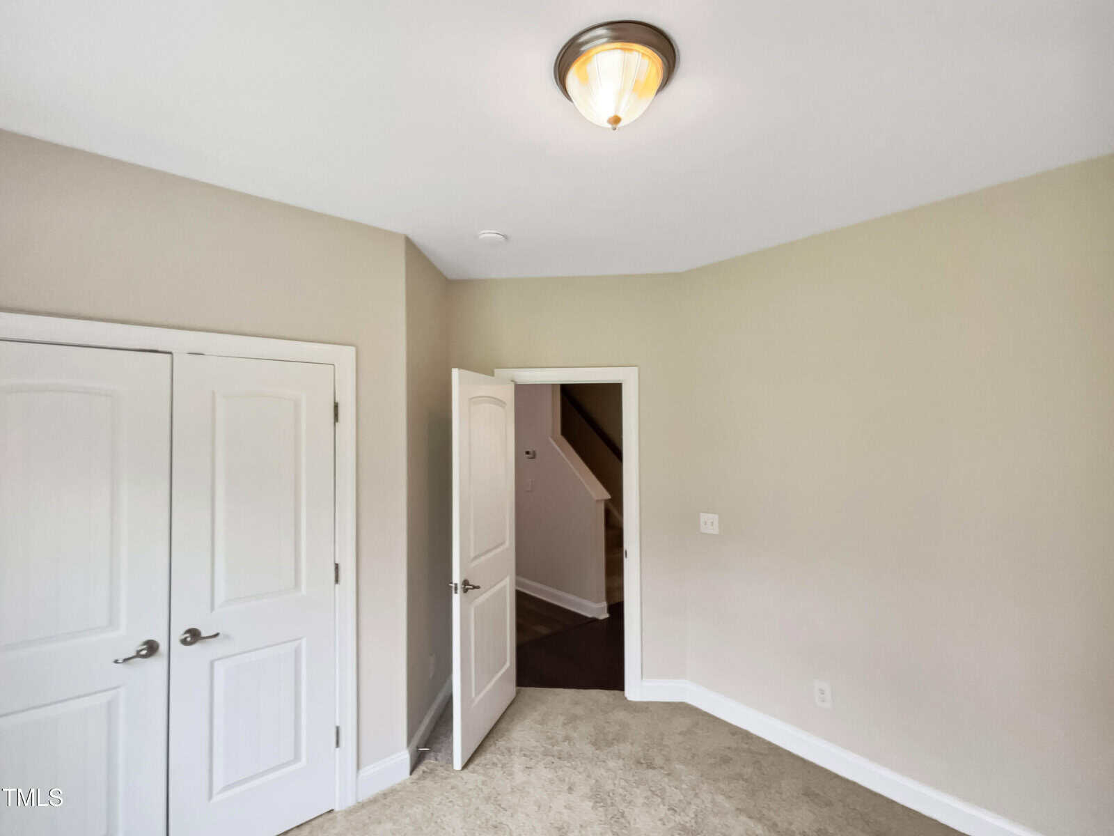 Empty room with beige walls and carpeted floor. A single light fixture is on the ceiling. There are two closed double doors on the left and an open door leading to a hallway in the center.