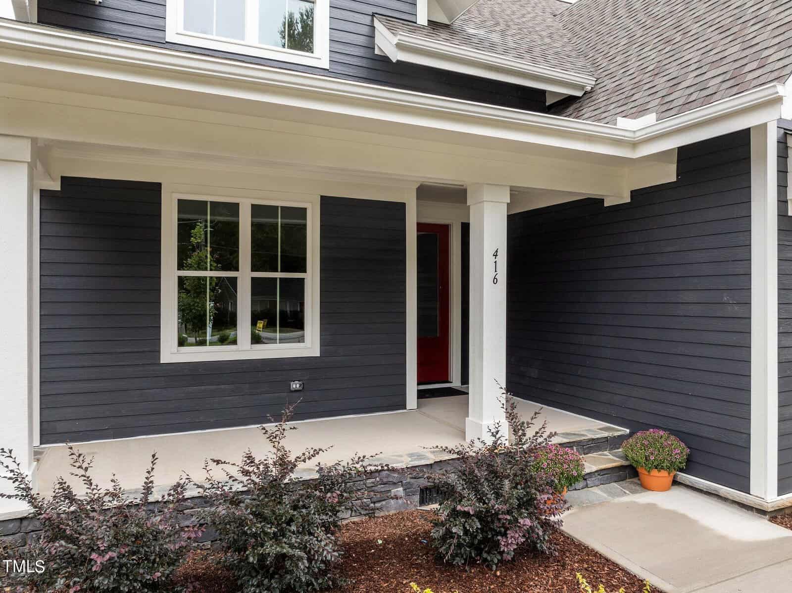 A modern house entrance with dark siding, a red front door, a covered porch, and a potted plant by the steps. Bushes and mulch line the edge of the porch.