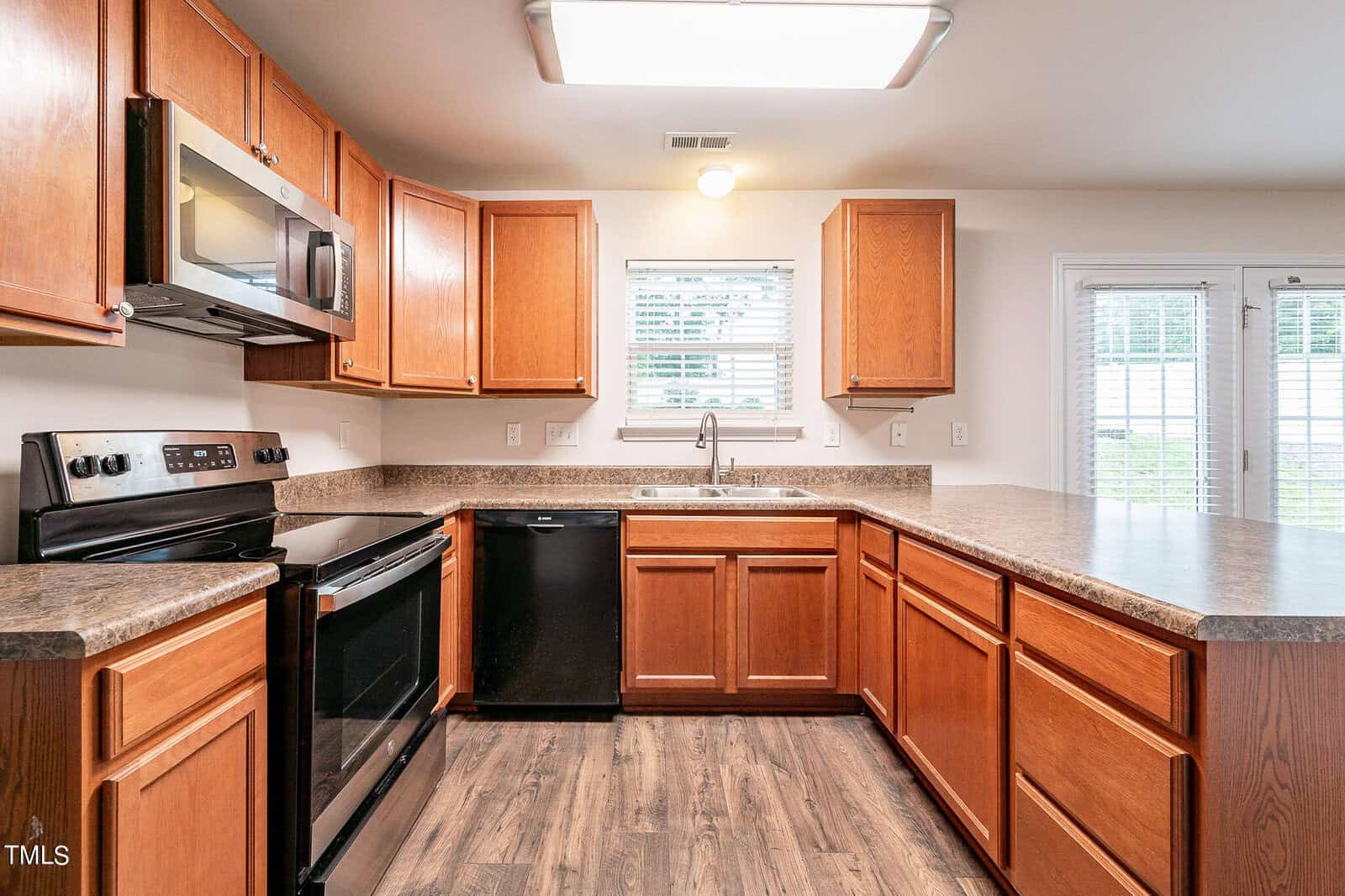 A spacious kitchen with wooden cabinets, stainless steel microwave, black oven, and dishwasher. The countertops are dark, and there is a window above the sink. Wood flooring and sliding glass doors with blinds are visible.