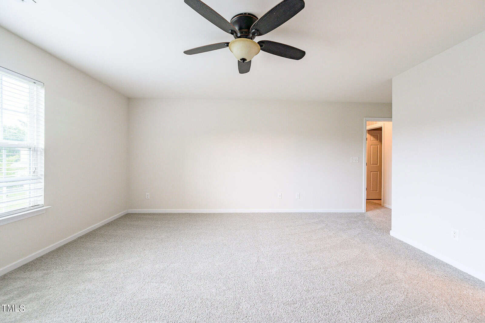 A spacious empty room with light gray carpet, white walls, and a large window on the left allowing natural light in. A ceiling fan with dark blades is centered on the ceiling, and an open door to the right leads to another area.