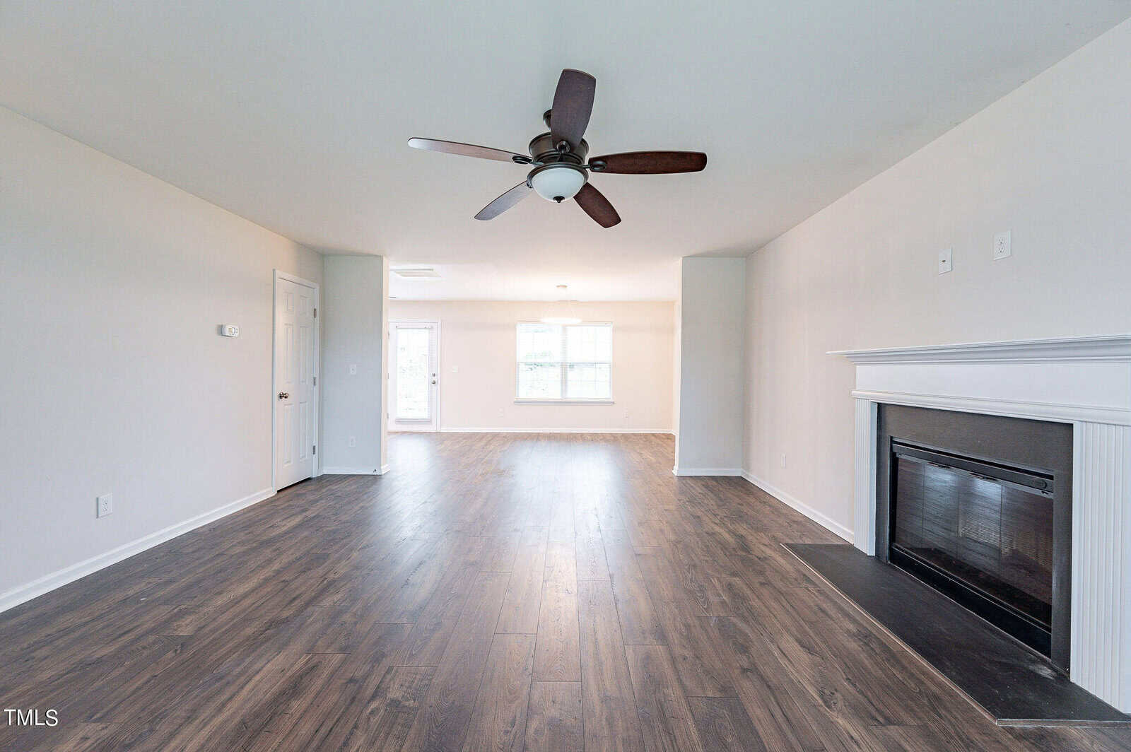 A spacious living room with light-colored walls and a dark wooden floor. A ceiling fan is mounted in the center. Natural light comes through two windows at the far end, and there is a fireplace on the right wall.
