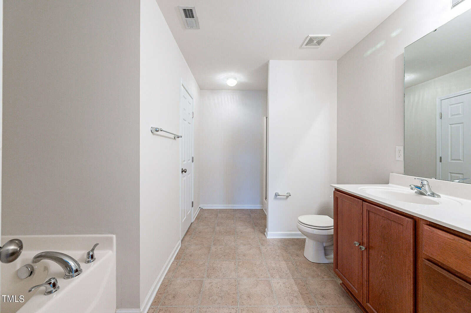 Spacious bathroom with light-colored tile flooring, featuring a large vanity with a mirror, a white toilet, and a bathtub with a chrome faucet. White walls and ample lighting enhance the clean, modern look.