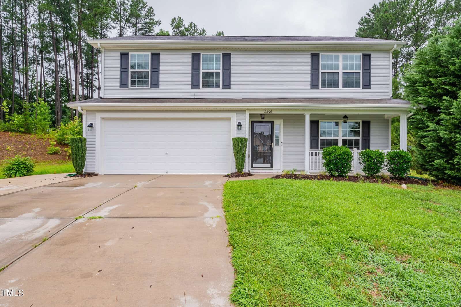 At 2206 Hyde Court, this two-story gray house in Creedmoor features white trim and black shutters. An attached two-car garage and concrete driveway lead to a front porch with pillars. Neatly trimmed bushes and a lawn surround the home, framed by tall trees in the background.