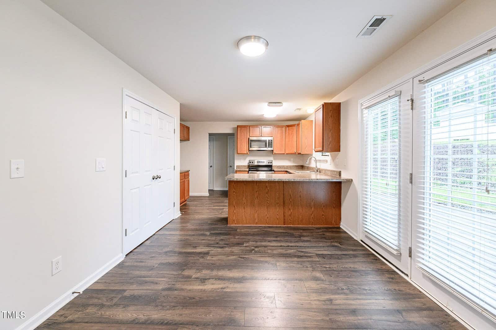 A bright, modern kitchen with wooden cabinets, stainless steel appliances, and granite countertops. Hardwood floors lead to a dining area with large windows and white blinds. A door opens to an outdoor space. Neutral walls and overhead lighting complete the room.
