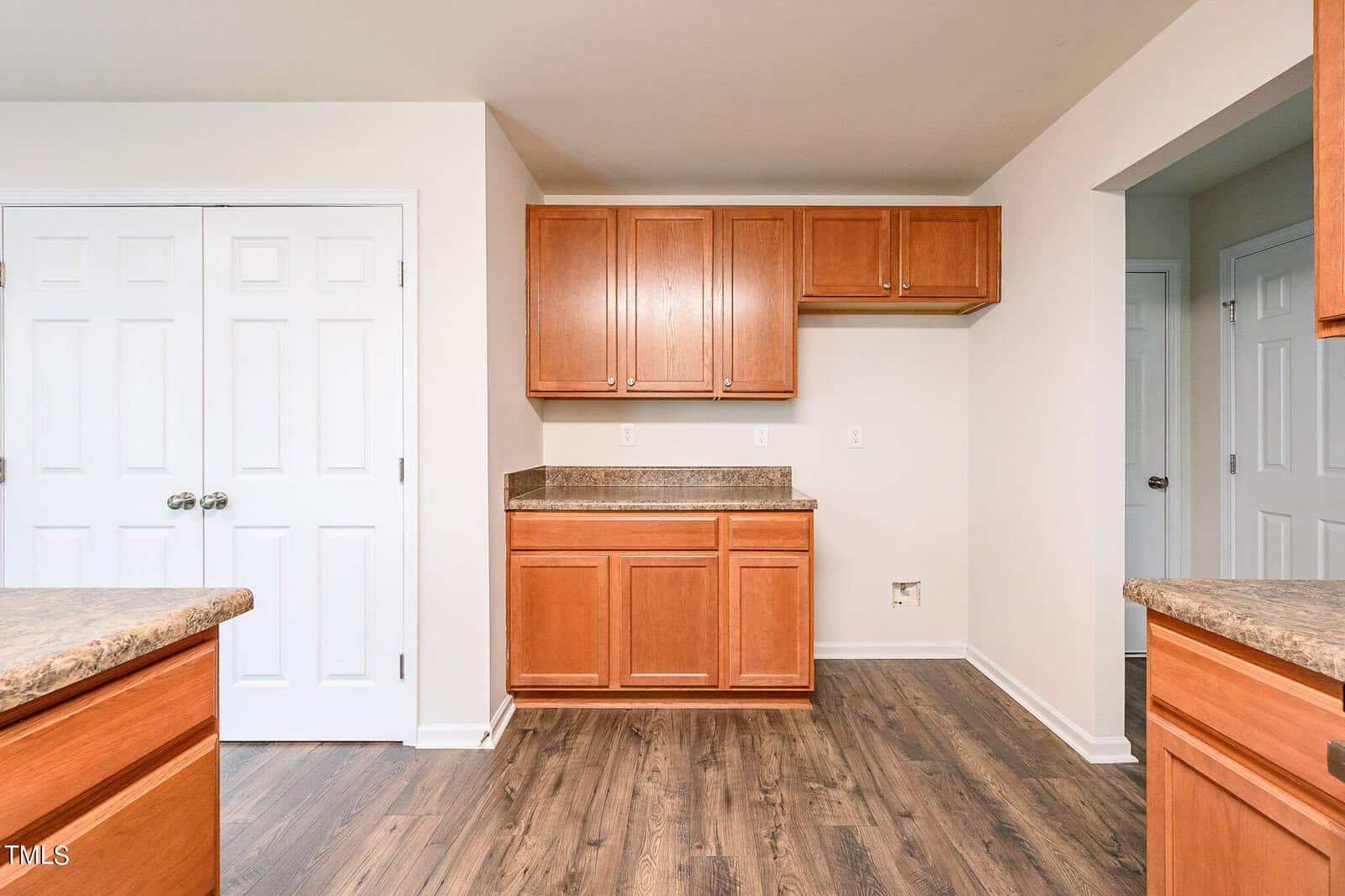Kitchen featuring wooden cabinets, light brown countertops, and wood flooring. There's a double-door closet on the left, and an open doorway on the right. The space is empty and well-lit, highlighting the clean, modern design.