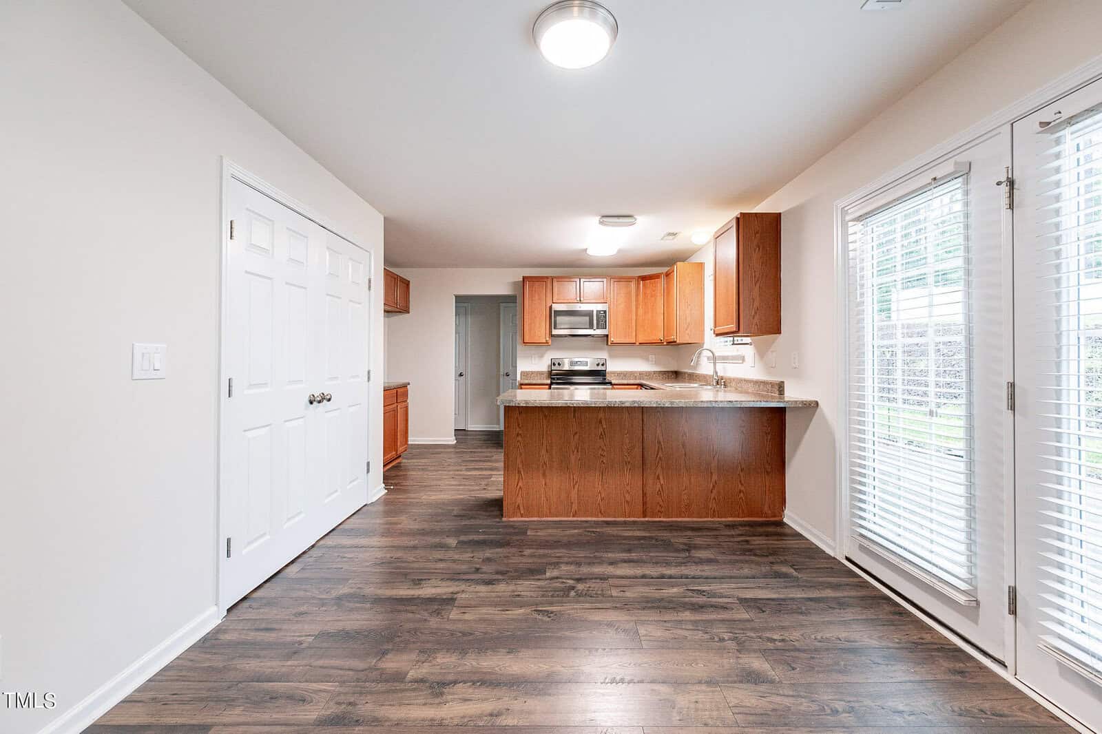 A modern kitchen with wooden cabinets, stainless steel appliances, and a central island. The room features a hardwood floor and sliding glass doors on the right, allowing natural light to brighten the space. A large closet is visible on the left wall.