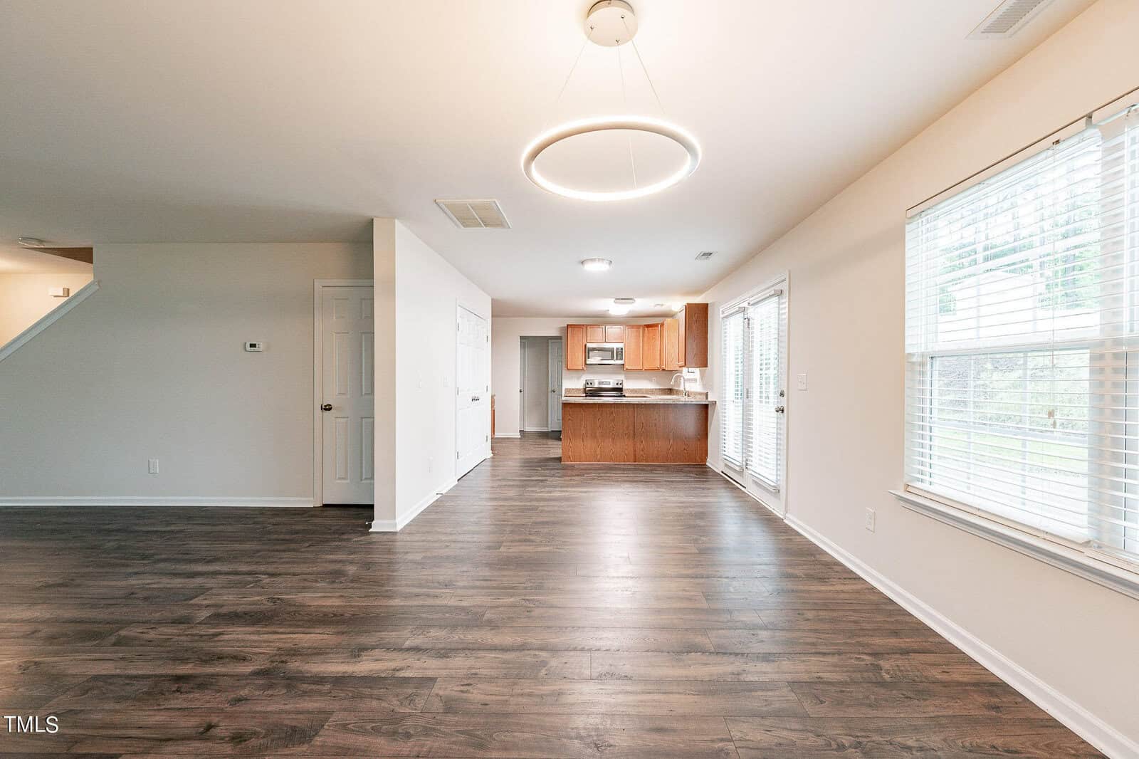 Spacious open-plan room with wood flooring and soft lighting. Kitchen area features wooden cabinets, stainless steel appliances, and a modern circular light fixture above. Large windows on the right let in natural light.