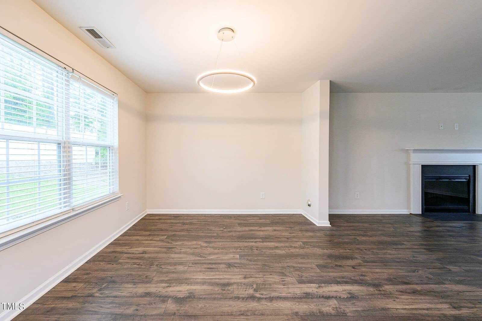 A spacious, empty room with wooden flooring, white walls, and a modern circular ceiling light. Large windows on the left let in natural light. A white fireplace is on the right with a flat wall adjacent.