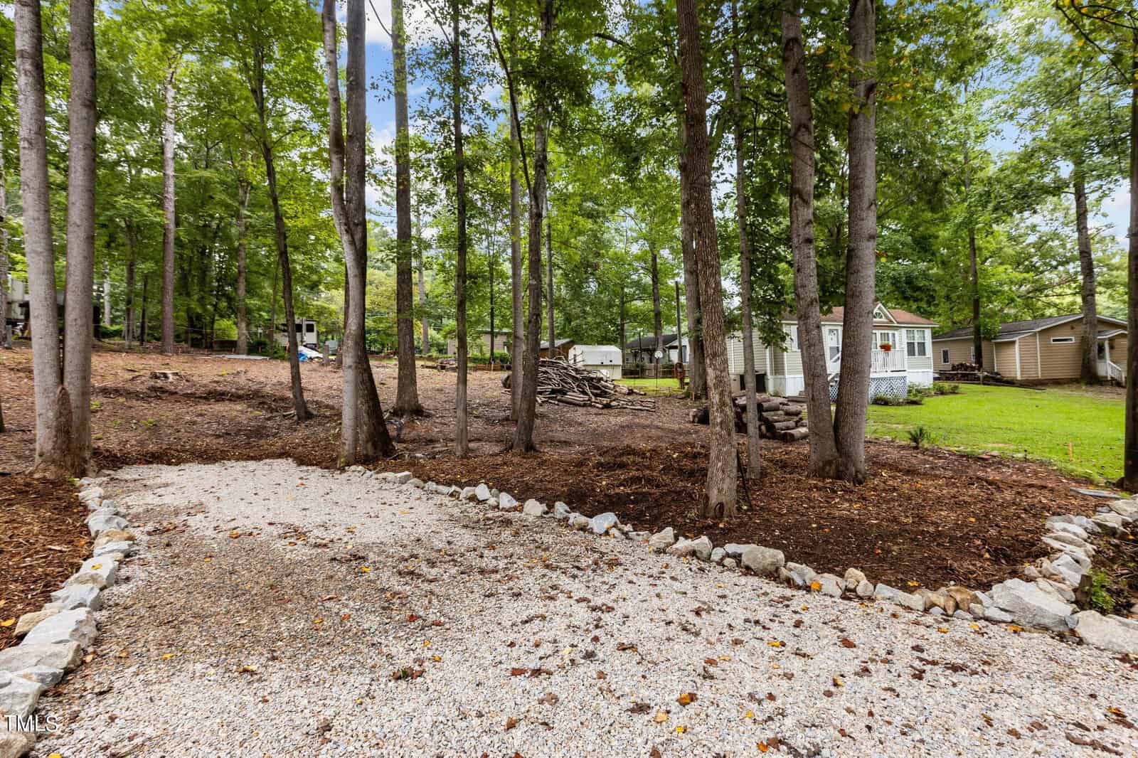 A gravel driveway bordered by rocks leads through a wooded area with tall trees toward small houses and cabins, with stacks of firewood and green grass visible in the background.