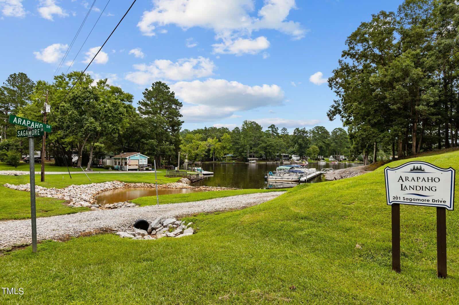 A tranquil marina with boats docked along a tree-lined shore, lush green grass, a street sign for Arapaho Street, and a white sign reading "Arapaho Landing, 201 Sagamore Drive" under a blue sky with clouds.