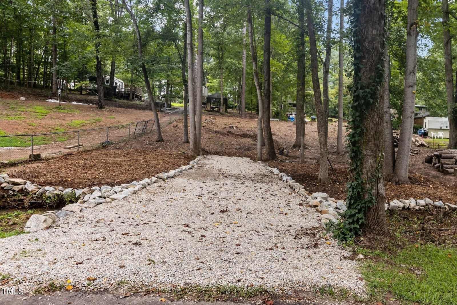 A gravel driveway lined with rocks leads uphill through a wooded area with tall trees and sparse grass, fenced areas, and a few sheds in the background.