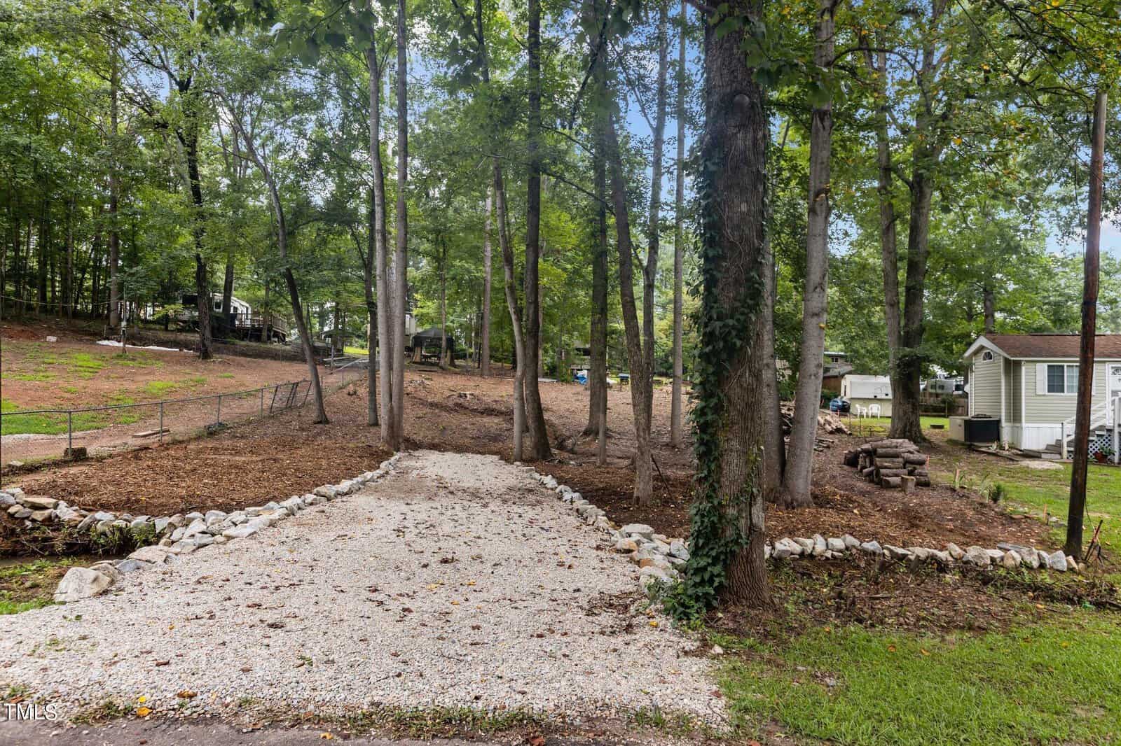 A gravel driveway bordered by rocks leads into a wooded area with tall trees and scattered leaves. To the right, there is a small light-colored house and wood piles. Other homes are visible in the background among the trees.
