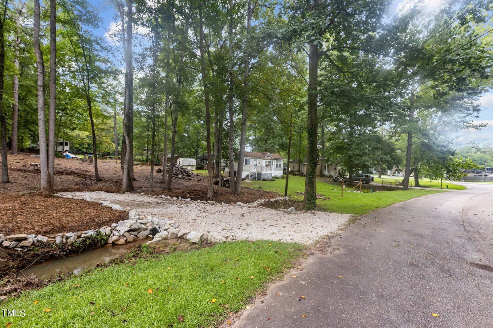 A gravel driveway leads through a wooded area toward a white house with a covered porch. There is a small rock-lined stream next to the driveway, and the scene is set on a quiet suburban street.