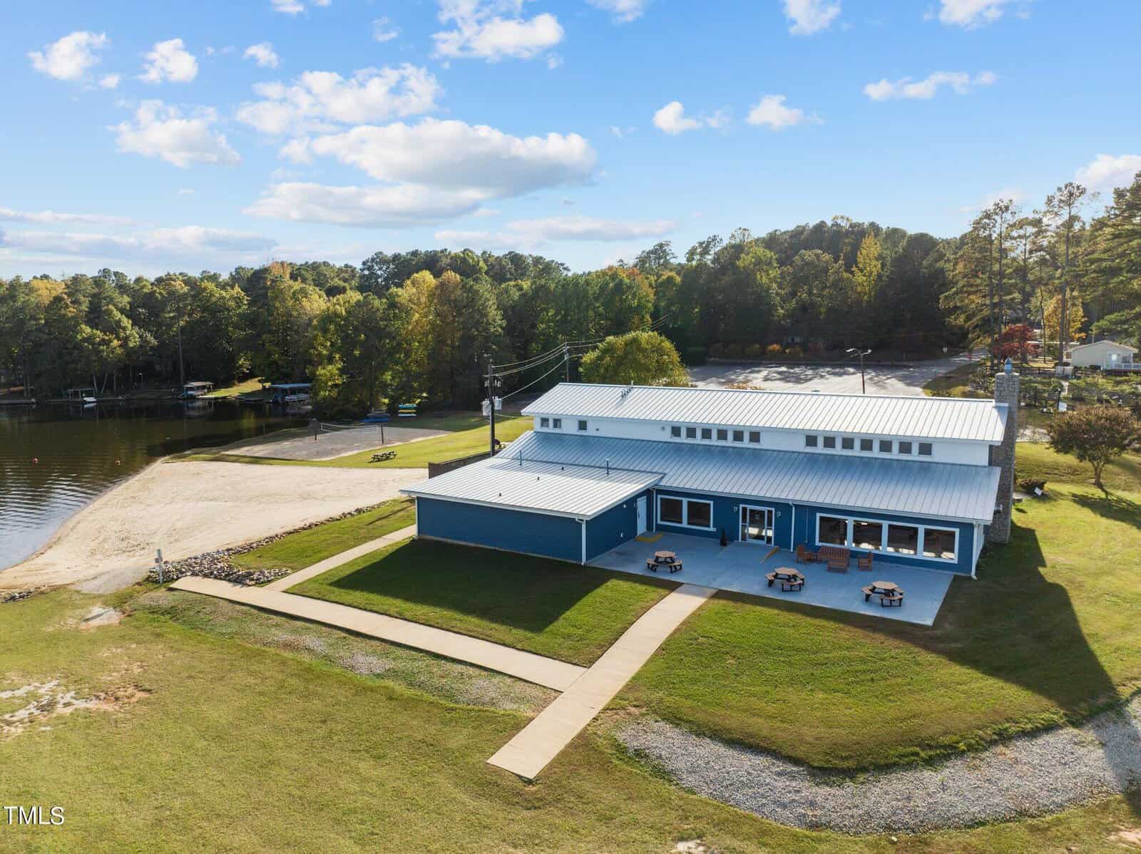 A large blue building with a metal roof sits by a sandy lakeshore, surrounded by grass and trees. Picnic tables are placed outside the building, and a paved walkway leads to the entrance. The sky is mostly clear with scattered clouds.