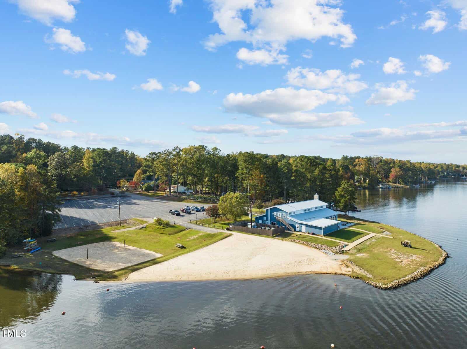 Aerial view of a small peninsula with a sandy beach, blue building, grassy areas, parking lot, and surrounded by water and trees under a partly cloudy sky.