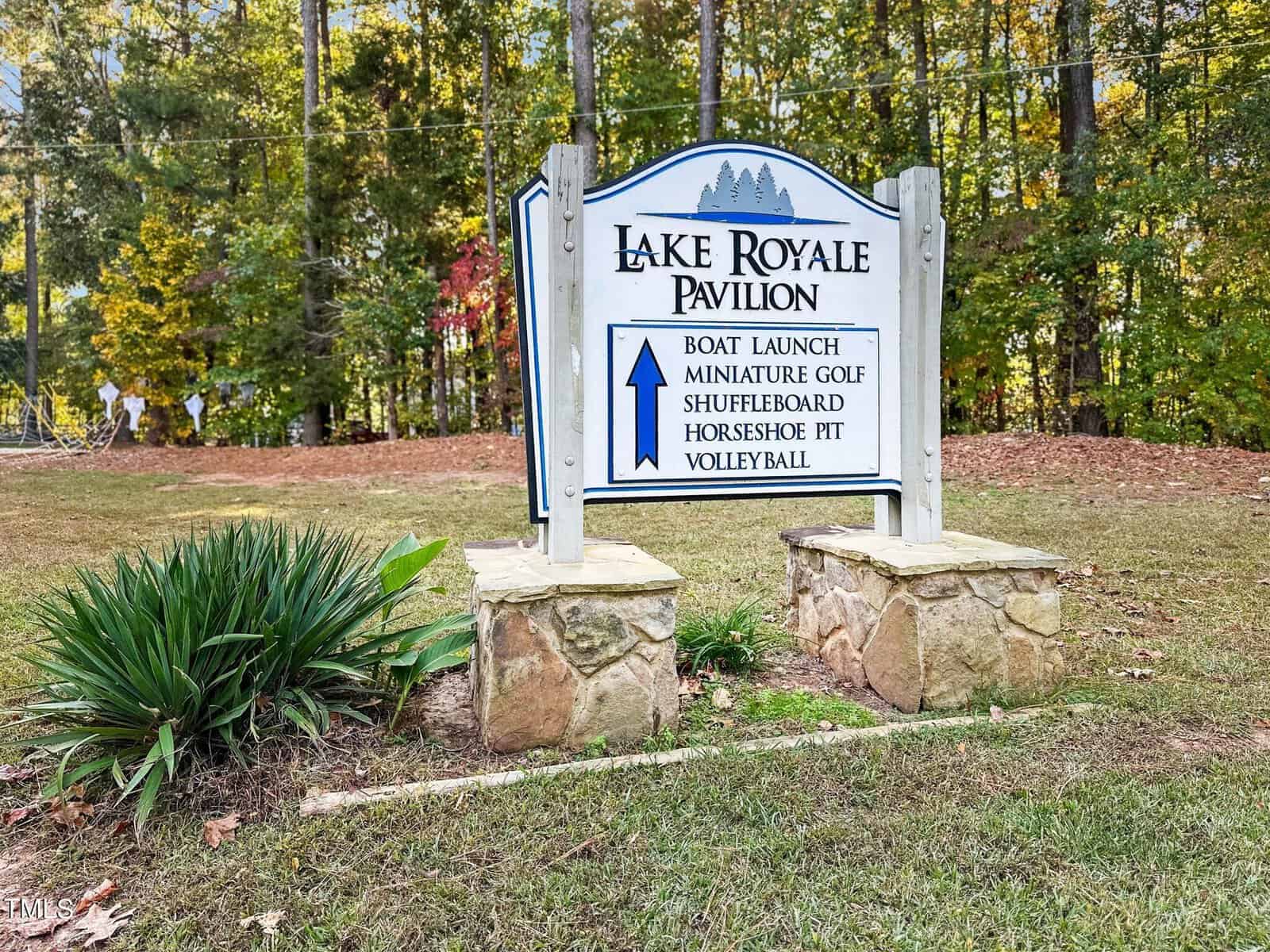 A white sign reads “Lake Royale Pavilion” and lists boat launch, miniature golf, shuffleboard, horseshoe pit, and volleyball, with an arrow pointing up. Surrounded by trees and grass.
