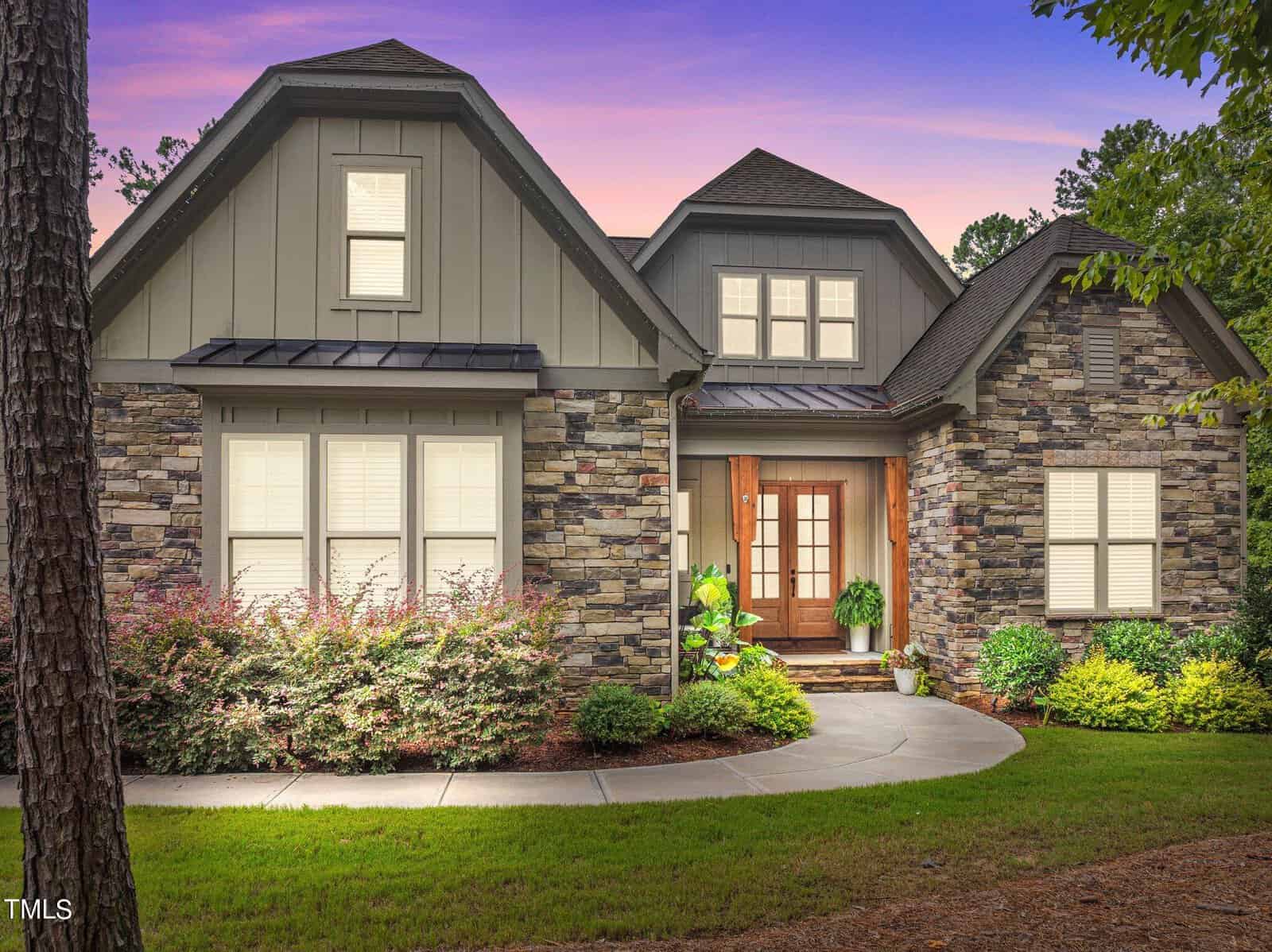 Located at 3593 Carole Court in Wake Forest, this modern house boasts a stone facade and multiple gables, surrounded by a manicured lawn and shrubs. It features a wooden front door with sidelights and a small front porch. The sky is a vibrant purple-pink, suggesting sunset or sunrise.