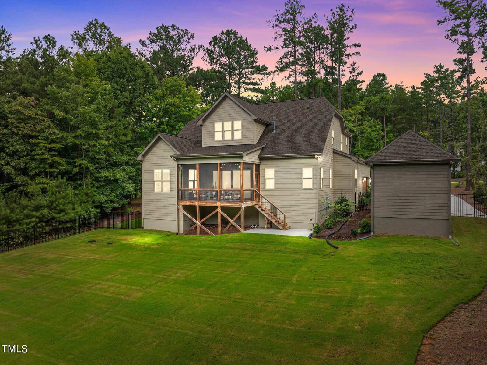 Back view of a modern two-story house with a large, manicured lawn. The home features a screened porch and is surrounded by tall trees under a purple and pink sunset sky. A separate garage is visible to the side.