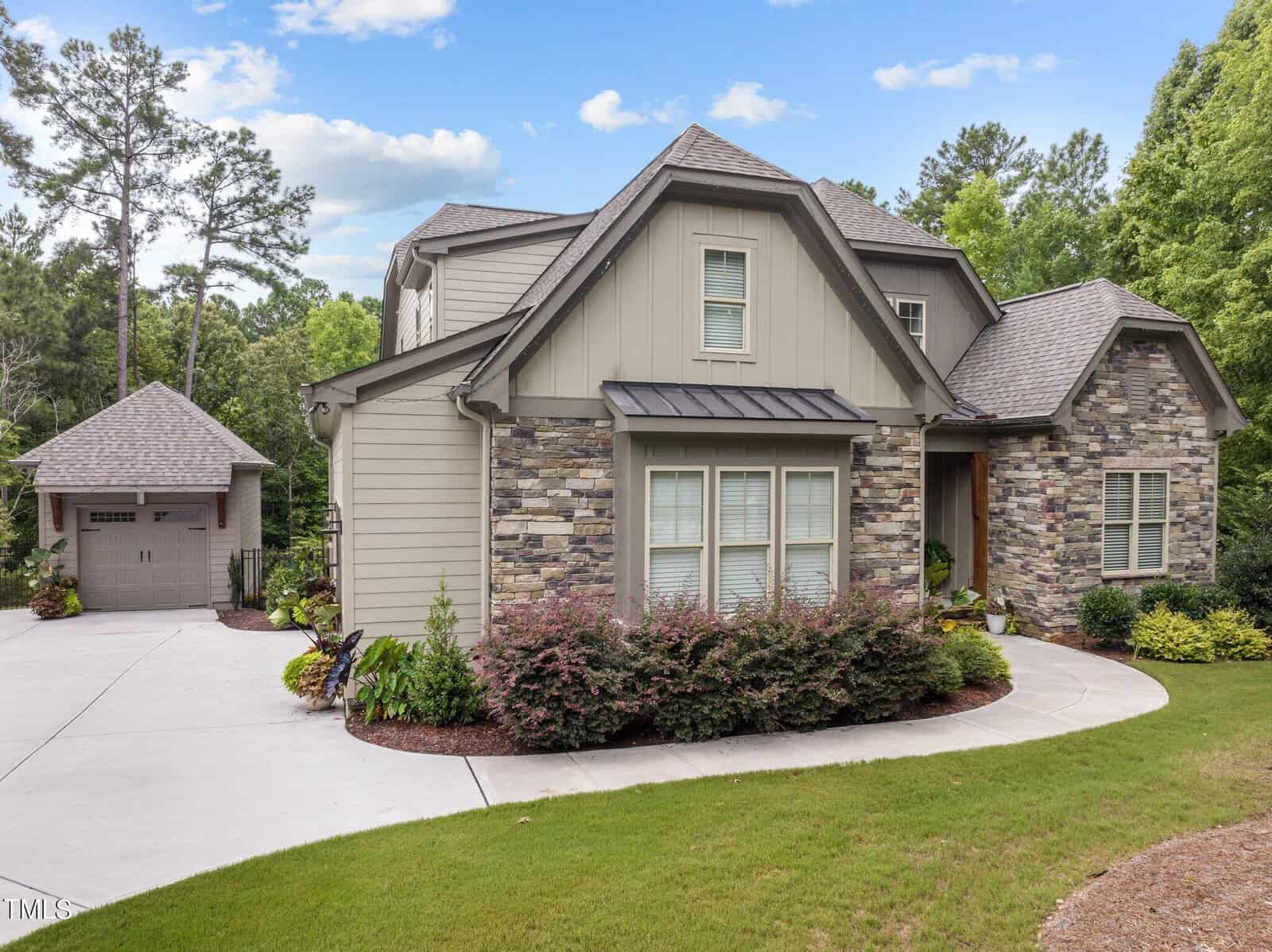 A modern house with stone and wood siding is surrounded by lush greenery. It features large windows and a steep roof. A detached garage is visible on the left, with a curved driveway leading to the entrance. The sky is partly cloudy.