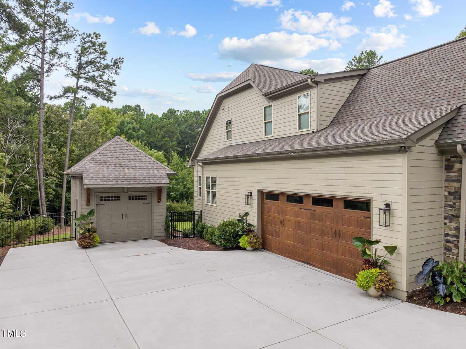 A modern house with beige siding, a two-car garage with wooden doors, and a separate smaller structure. The driveway is expansive and clean. The property is surrounded by lush green trees under a partly cloudy sky.