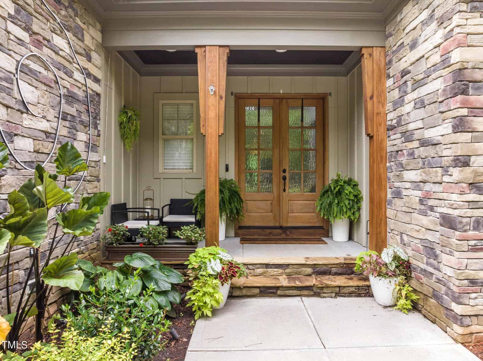 A welcoming front porch with wooden double doors framed by stone walls and wooden beams. Potted plants with lush greenery and a small seating area with black chairs add to the cozy atmosphere. A modern circular metal sculpture adorns the stone wall.