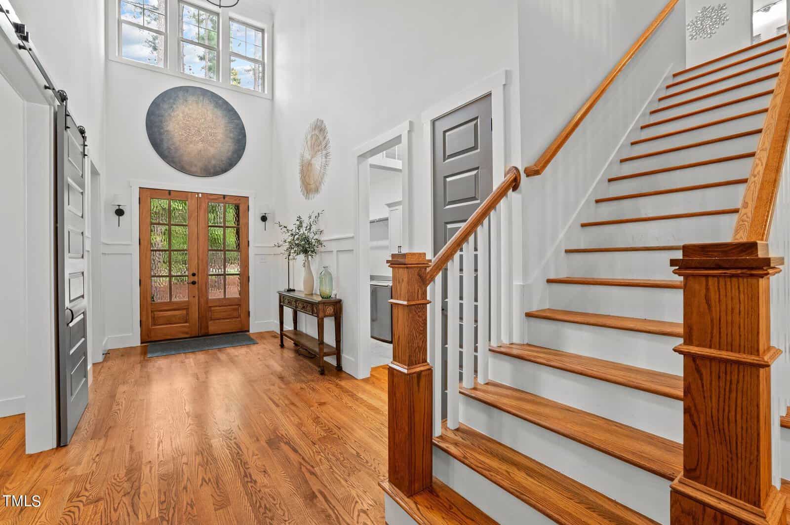 A bright, airy foyer with wooden floors and stairs. It features a large front door with glass panels, decorative wall art, a console table with a plant, and a staircase with white railing. High windows let in natural light.