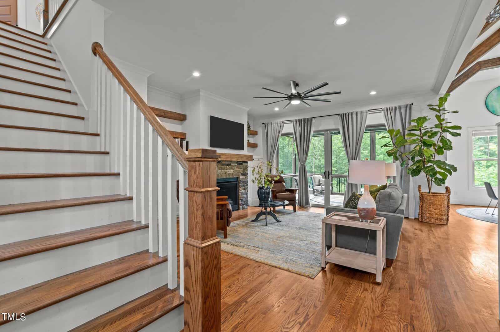 A spacious, elegant living room with hardwood floors and a large staircase. There's a stone fireplace, a modern ceiling fan, and large windows with curtains offering a view of greenery. A cozy seating area with a rug and plants completes the space.