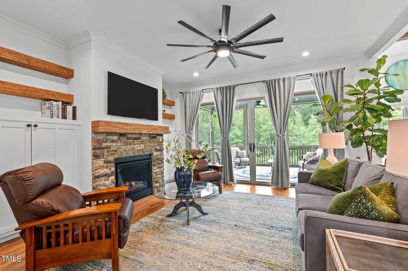 A cozy living room with a stone fireplace and wooden mantel, featuring a mounted TV above. The room has a sofa, two chairs, a glass coffee table, and a large window with curtains overlooking a lush outdoor area. A ceiling fan hangs above.