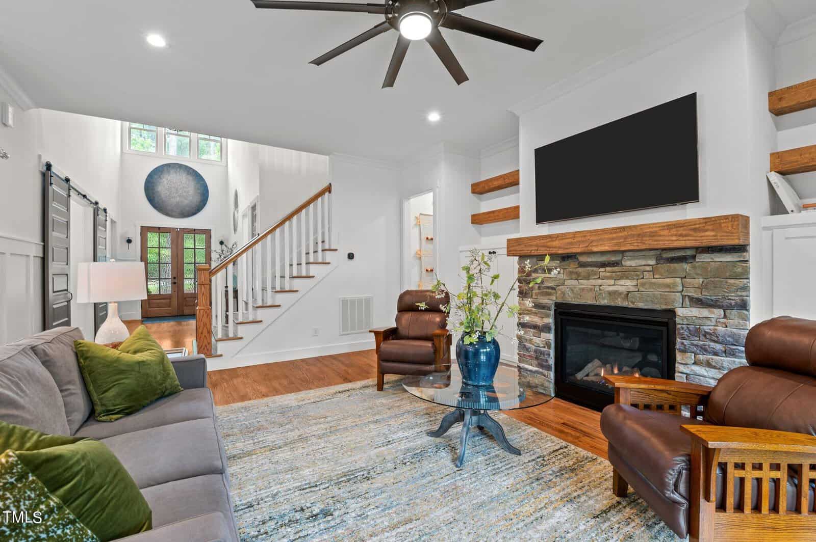 Modern living room with wooden floors, a gray sofa, and two brown leather armchairs. A stone fireplace features a TV above it. A large rug covers part of the floor. Wooden shelves are on the wall, and a ceiling fan hangs from the ceiling. Stairs in the background.
