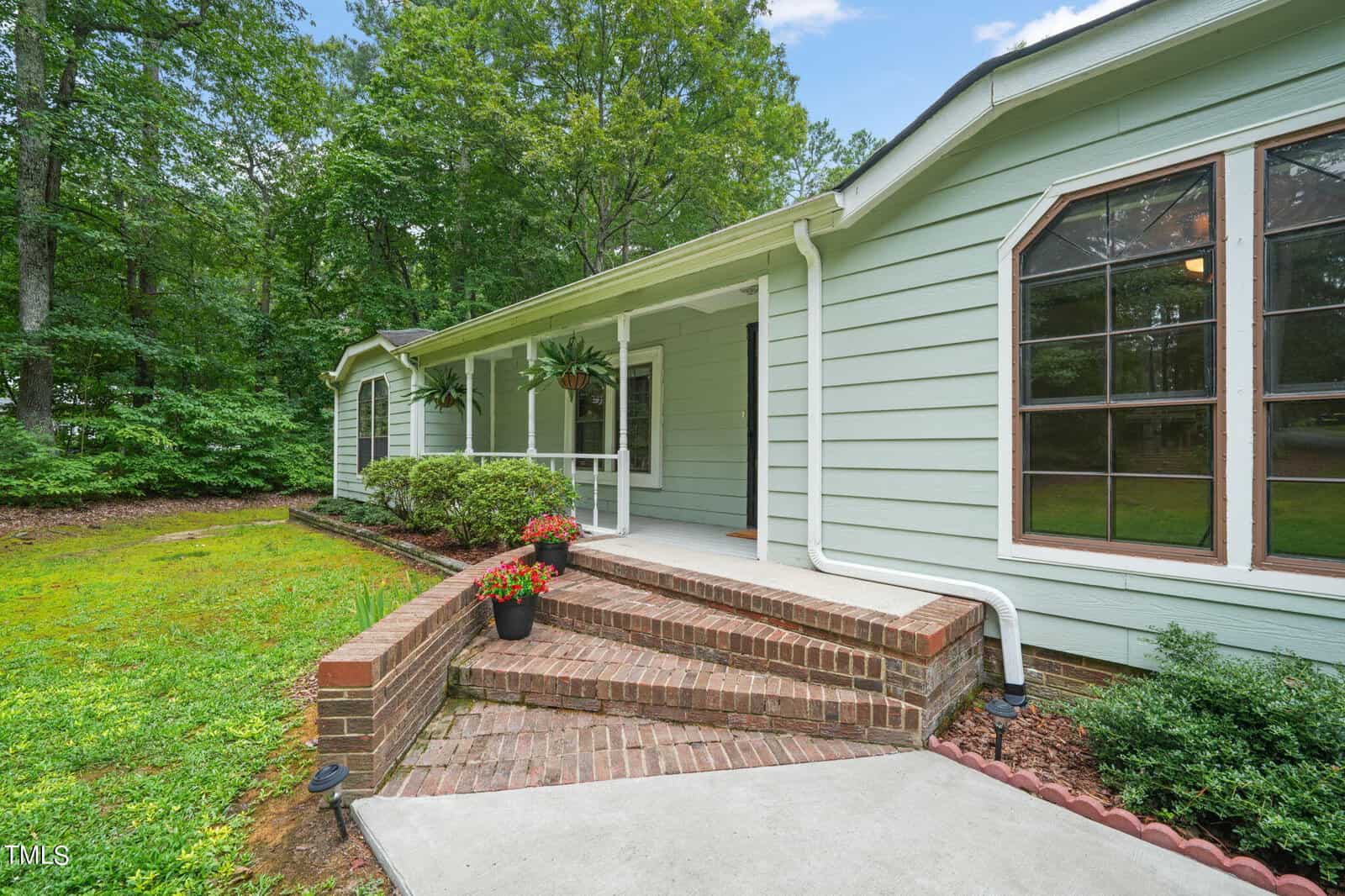 A light green single-story house with large windows, surrounded by lush greenery. The home features a brick walkway and steps leading to a covered entrance. Potted plants decorate the steps, enhancing the welcoming atmosphere.