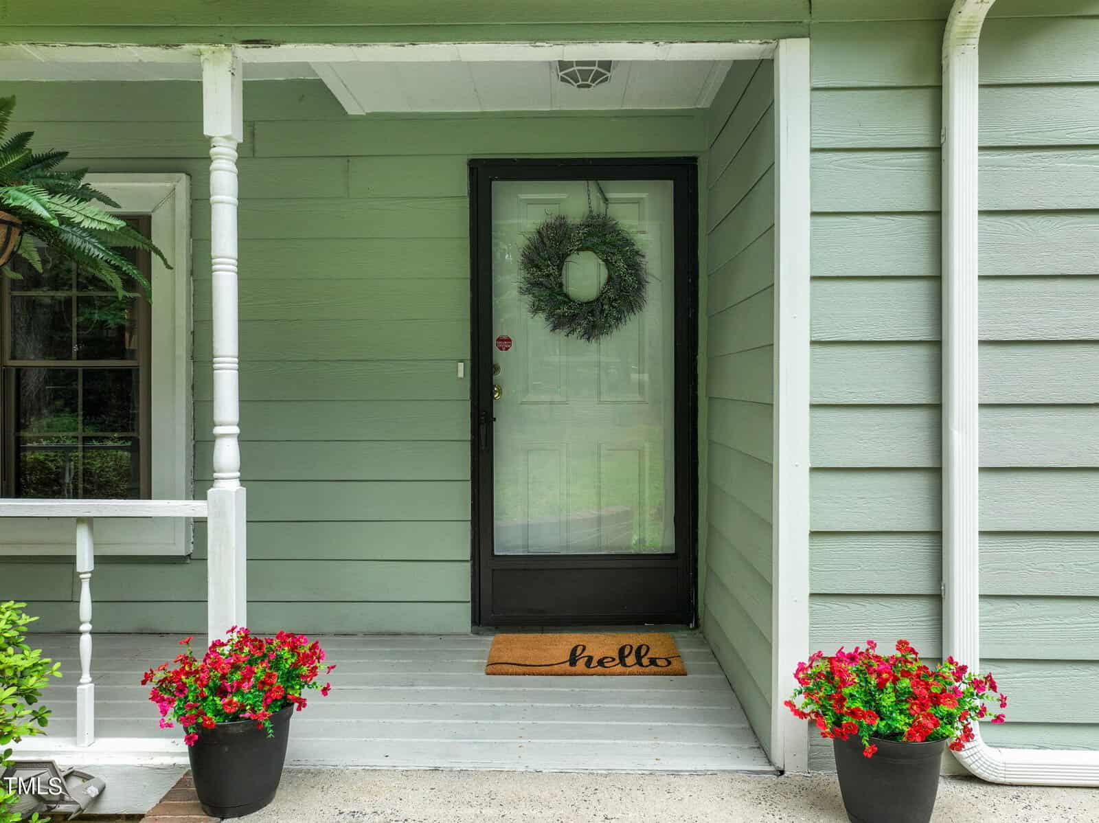 A welcoming front porch with a green exterior wall and white door featuring a wreath. A "hello" doormat lies on the floor. Red flowers in pots are placed on the steps, and a fern hangs from the porch ceiling.