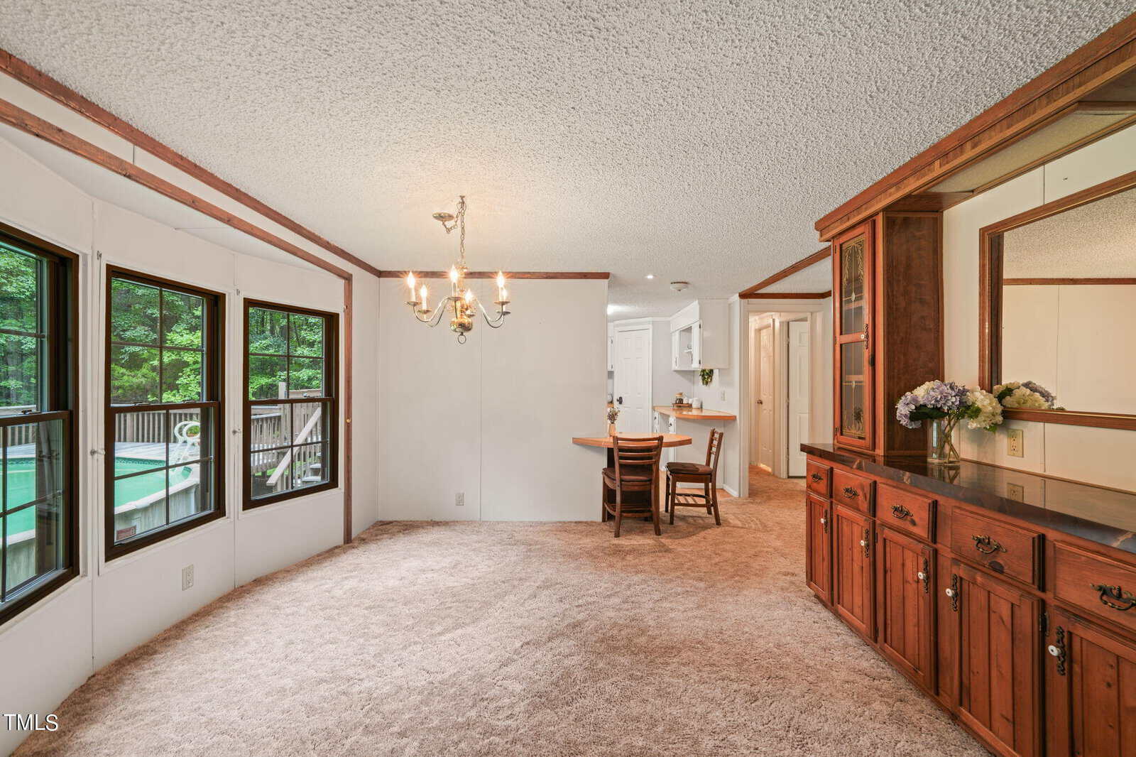 Dining area with large windows, beige carpet, and chandelier. To the right, a wooden cabinet with a mirror and floral arrangement. In the background, a wooden dining table with chairs is visible, adjacent to a bright kitchen.