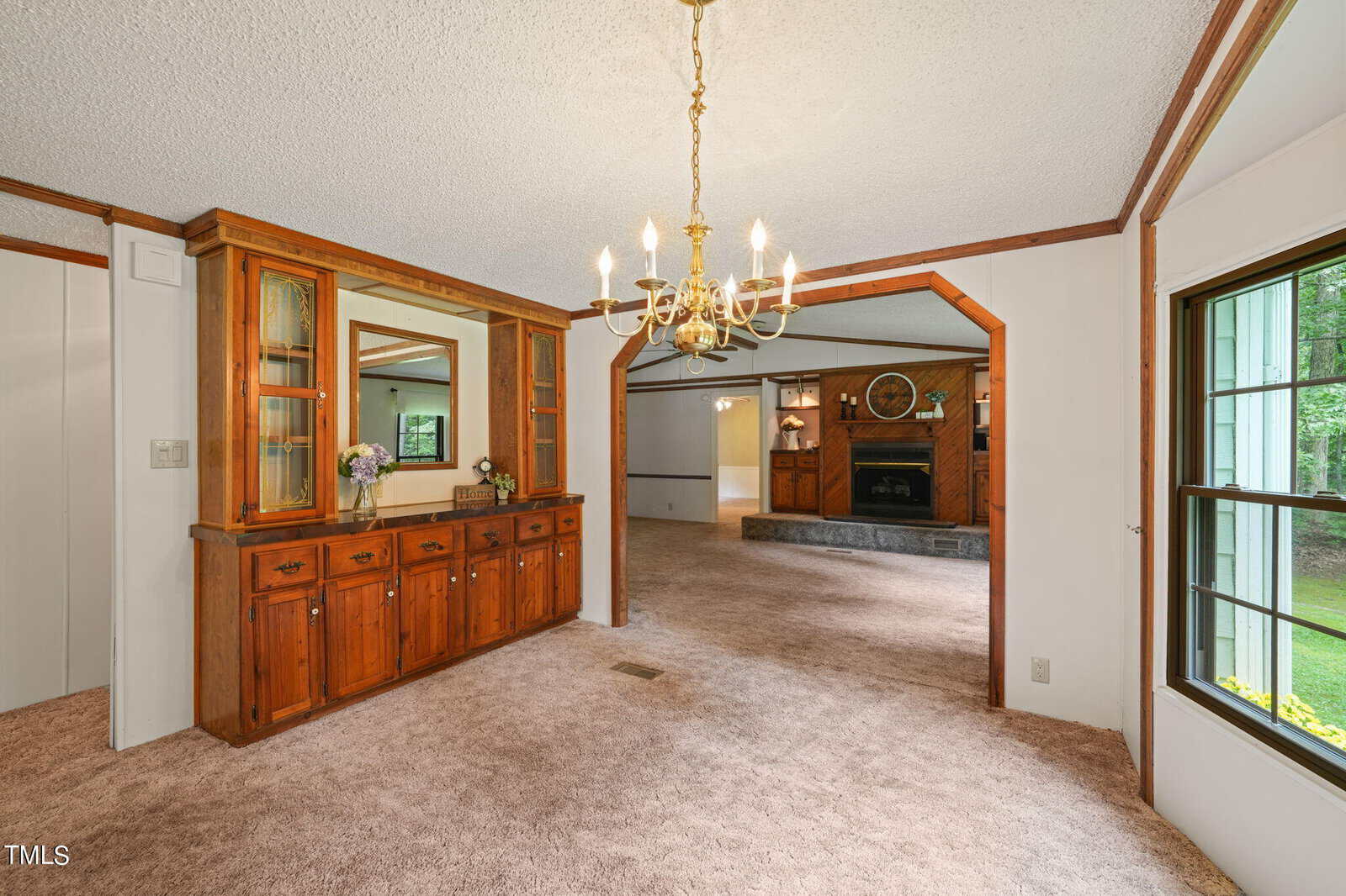 A cozy interior featuring a carpeted living room with a chandelier, wooden built-in cabinets, and a large mirror. A spacious adjacent room with a stone fireplace and decorative clock is visible. Large windows let in natural light.