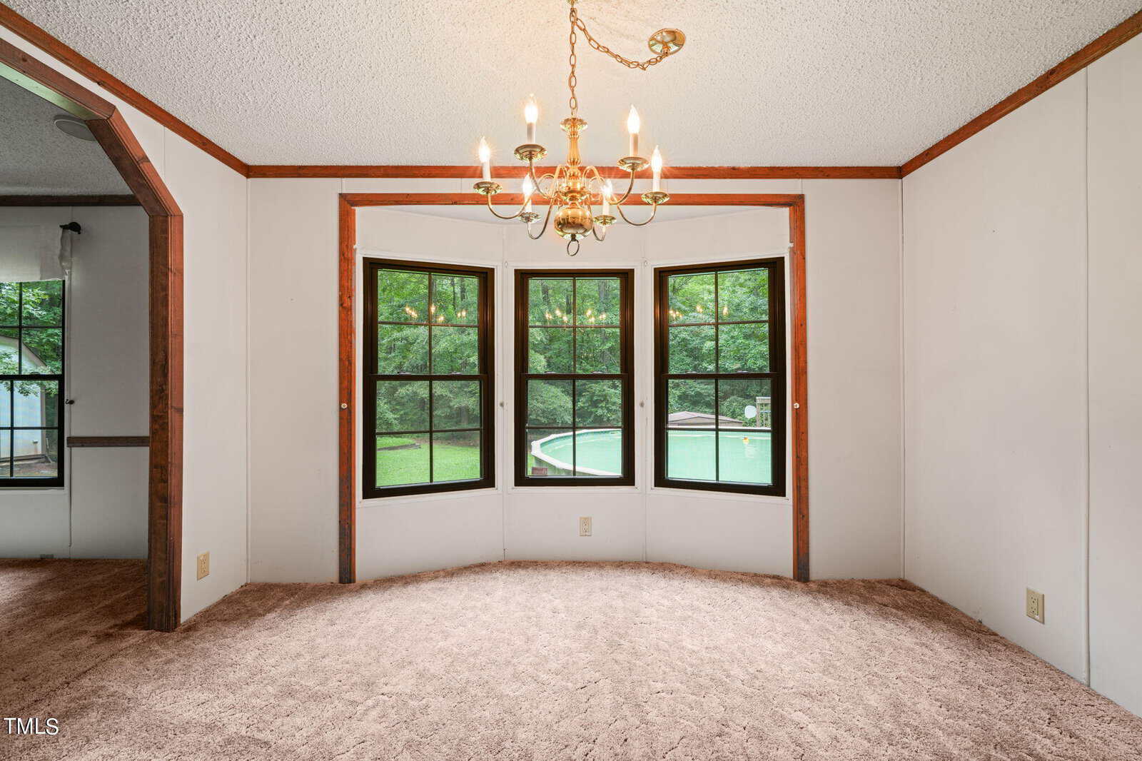 A dining room with beige carpet and white walls, featuring a gold chandelier. Three large black-framed windows overlook a green yard with a swimming pool. An arched doorway leads to an adjacent room with a curtained window.