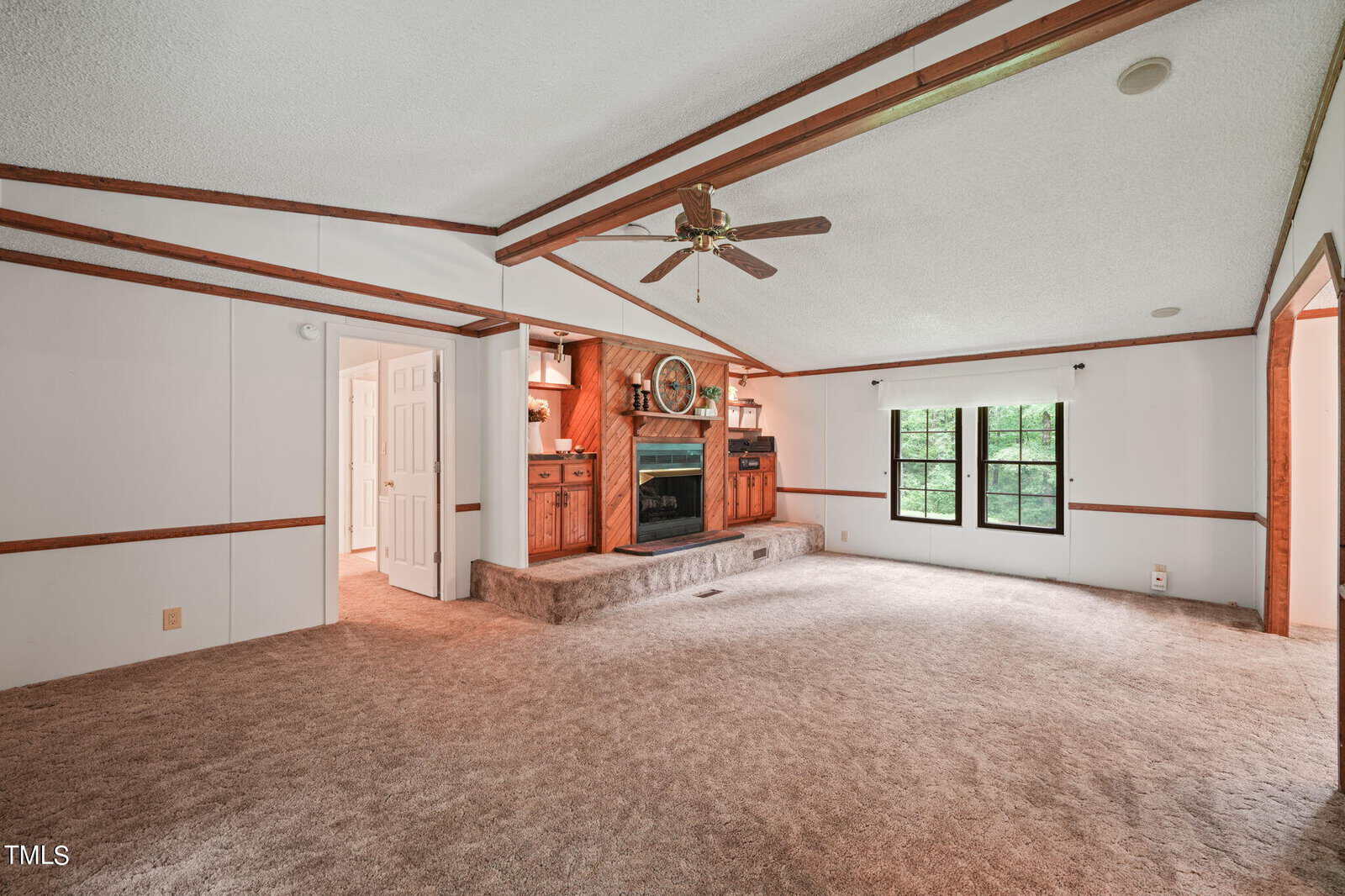 Spacious living room with carpeted floor, wood-paneled walls, and a ceiling fan. Features a large fireplace with a decorative mantel and mirror. Two windows allow natural light in, and an open doorway leads to another room.