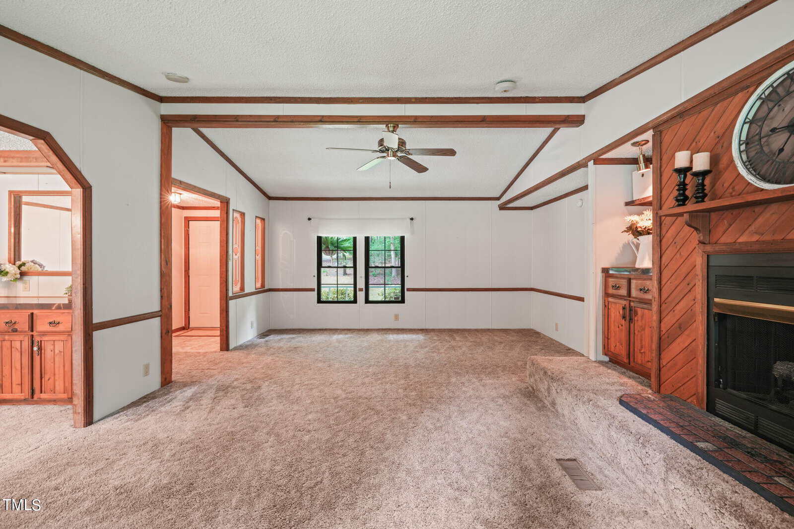A cozy living room with a beige carpet, white walls, and wooden beam accents. It features a ceiling fan, a large window with greenery visible outside, and a fireplace on the right with a mantel and decorative items. A glimpse of the kitchen is on the left.