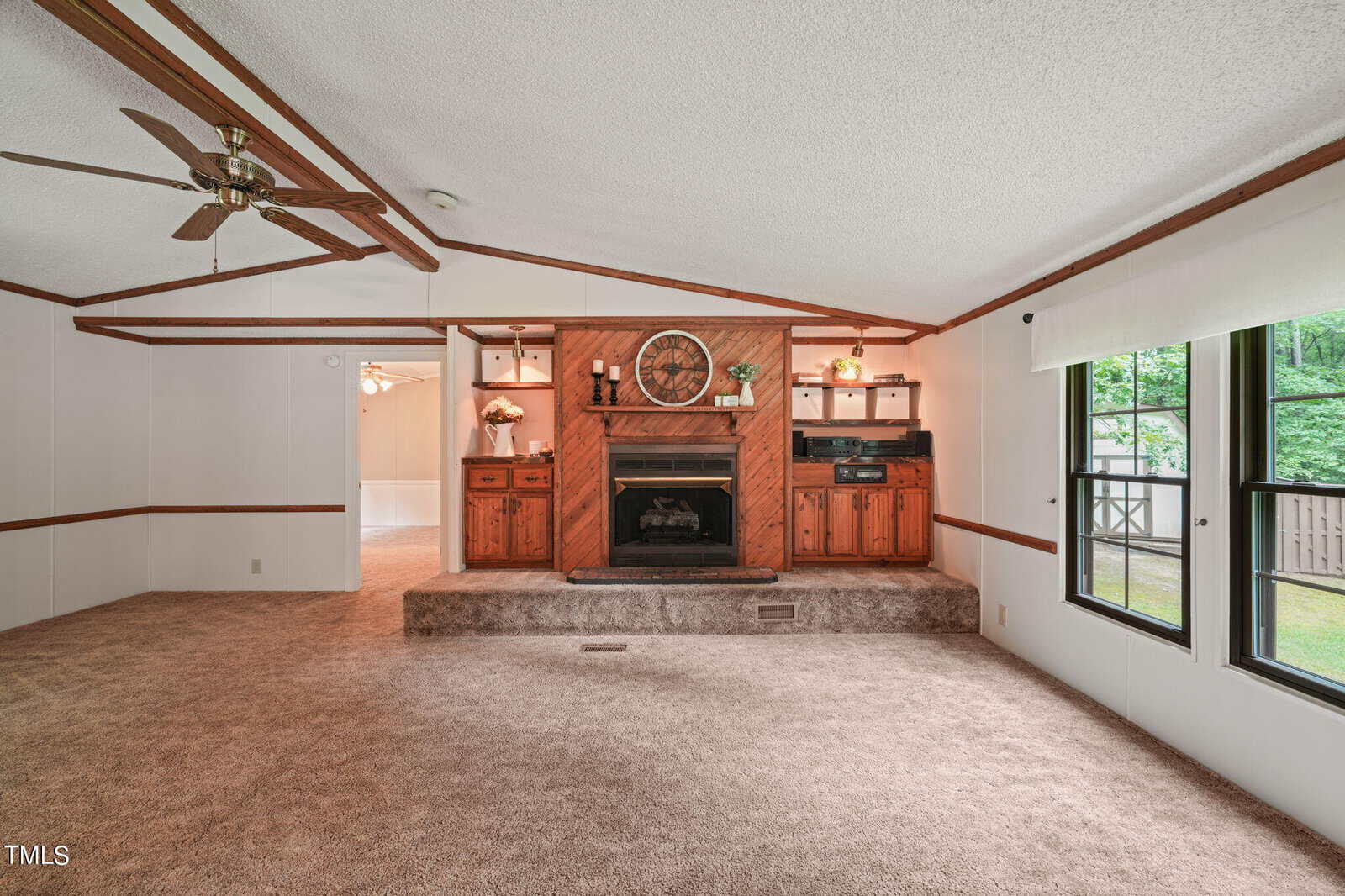A spacious living room with carpeted floors and a wooden accent wall featuring a fireplace. Above the fireplace is a decorative mirror and built-in shelves. Large windows on the right let in natural light. Ceiling fans are mounted on the white ceiling.