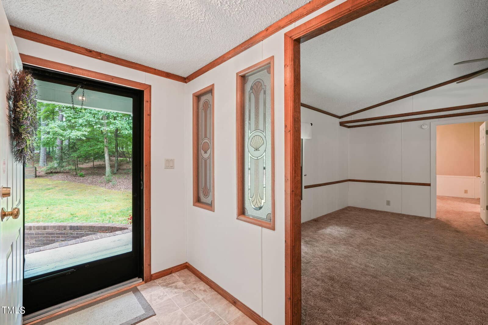 A bright entryway with light-colored walls and decorative glass windows. The door is open, showing a grassy outdoor area with trees. The adjacent room has carpeted floors and wood trim.