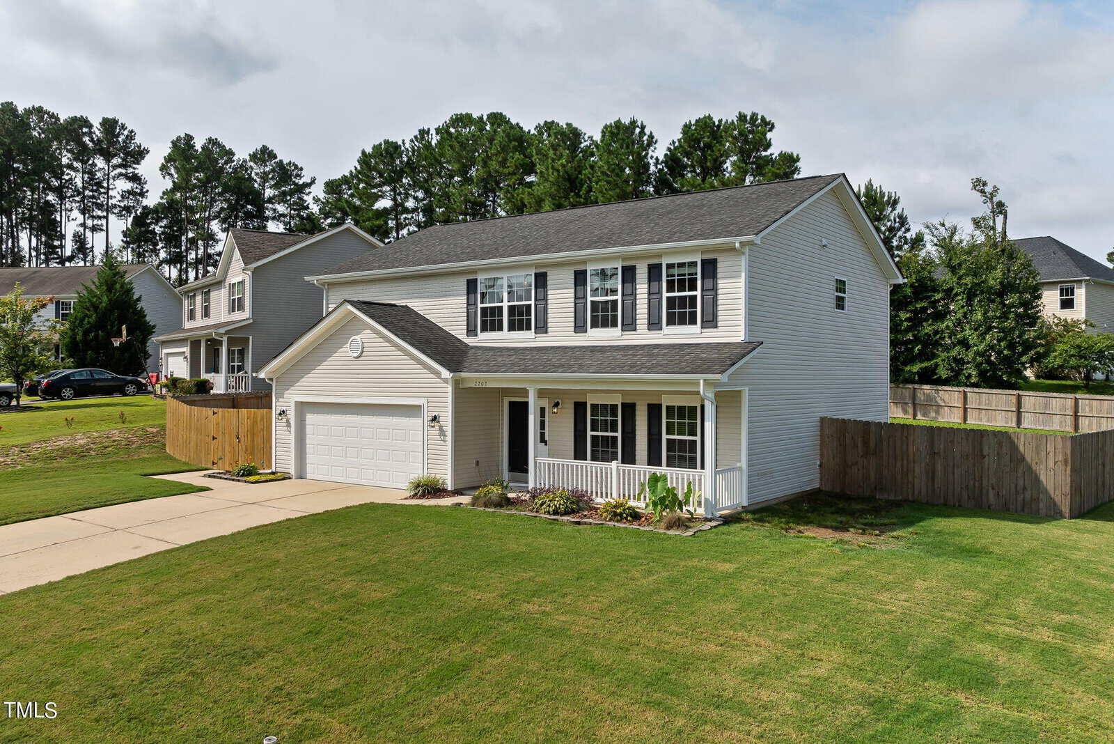 A two-story white house with grey roof shingles, a double garage, and a well-maintained lawn. It is surrounded by a wooden fence and tall trees in the background. The sky is partly cloudy.