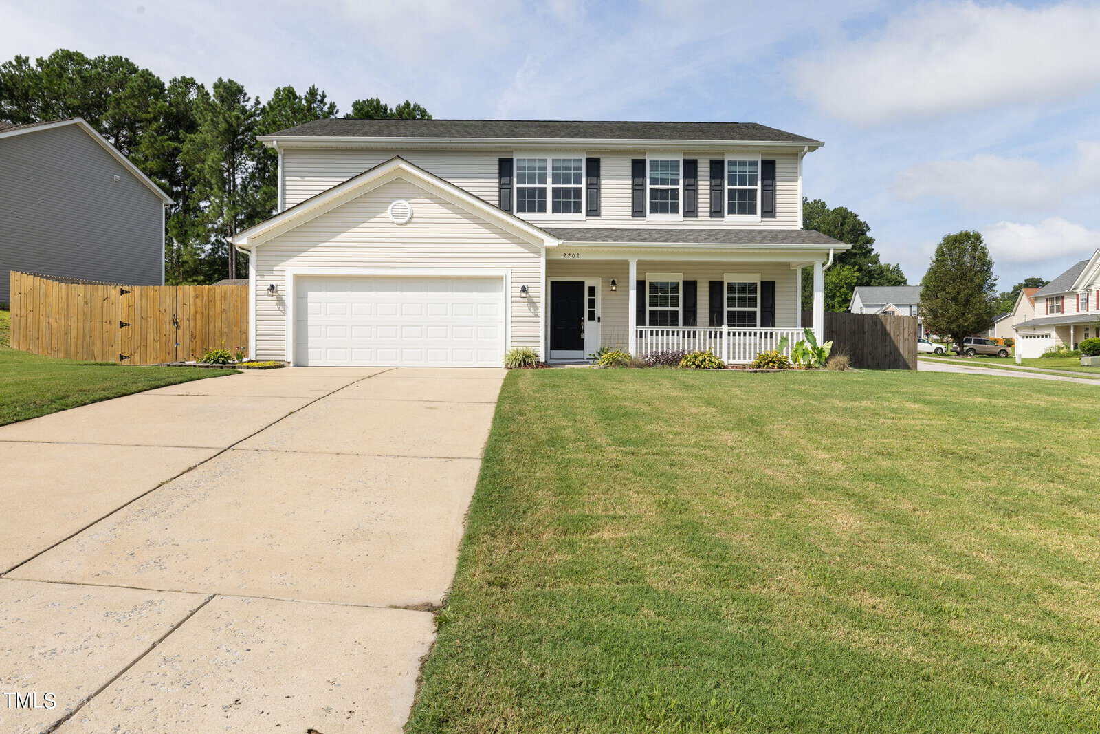 A two-story suburban house with white siding and a black roof, featuring a double garage and a front porch. The driveway leads to a well-maintained lawn, and the neighborhood has similar homes. Trees are visible in the background.