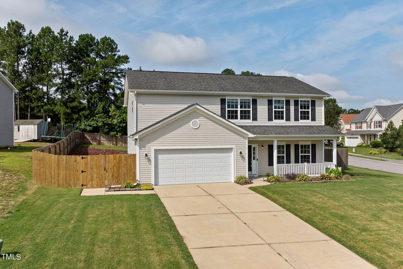 Two-story suburban house with a gray exterior, black shutters, and a double garage. It's set on a green lawn with a wooden fence in the backyard. The sky is blue with a few clouds.